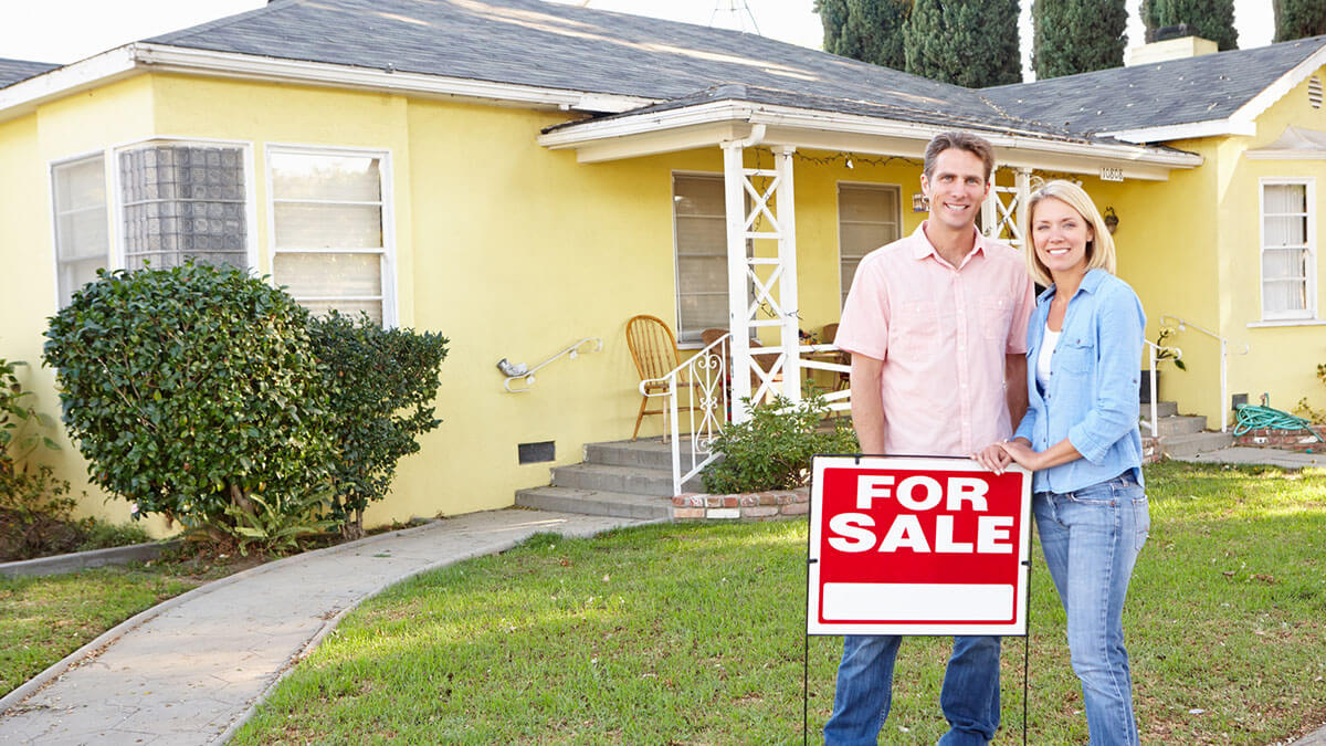 couple standing behind a for sale sign with their house that is for sale in the background