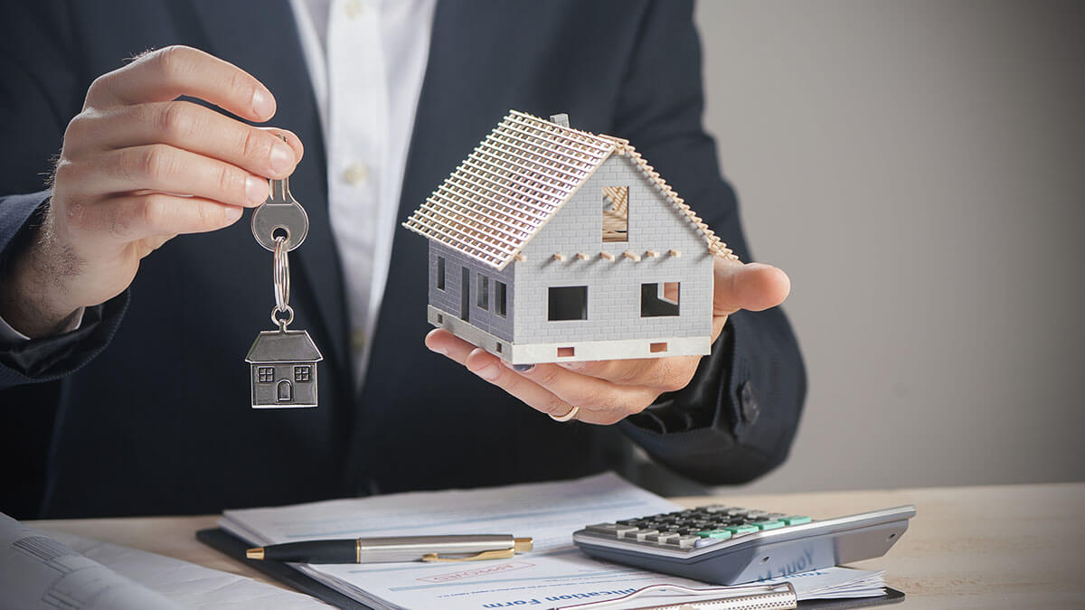 realtor holding miniature home and house keys with calculator, contract and pen on the table