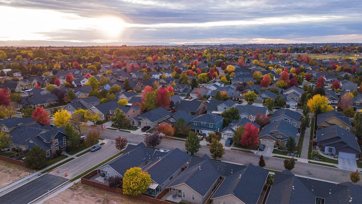 Aerial view of a suburban neighborhood during autumn, showcasing a variety of houses with colorful trees in shades of red, orange, and yellow, streets with parked cars, and a dramatic sunset in the background