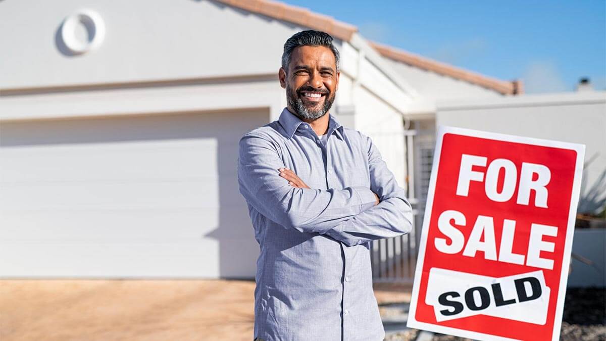 person standing in front of the house they have just sold with a sign on their right